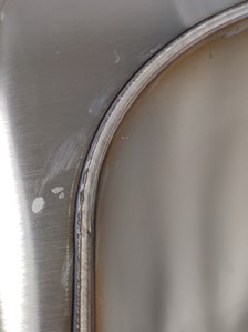 Kiiltävää metallia, jossa keskellä pystysuuntainen, ylhäällä oikealle kaartuva hitsaussauma.